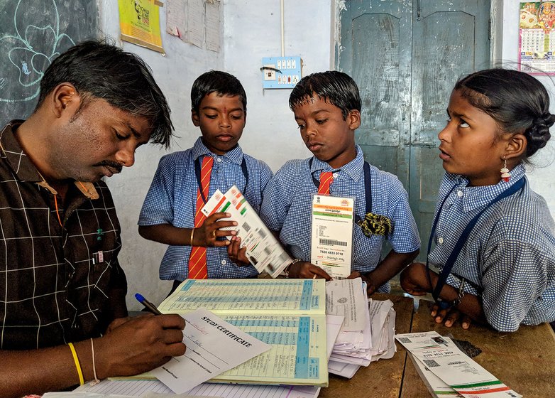 A man writing at a desk in a classroom surrounded by two young boys and a girl