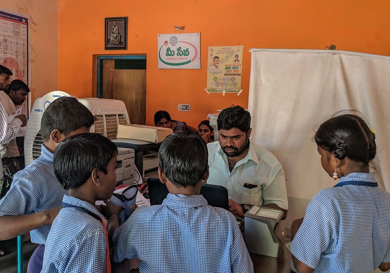 A man sitting at an office desk surrounded by three young boys and a girl wearing school uniforms